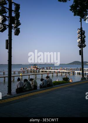 Wuhan, Cina - 16 giugno 2025: Persone che si rilassano su gradini di legno accanto a una passeggiata lastricata sul lungomare con pavimentazione tattile vicino a un lago. L'immagine presenta una Foto Stock