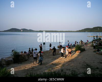 Wuhan, Cina - 16 giugno 2025: Persone che camminano e si rilassano su una spiaggia sabbiosa con grandi rocce vicino a un lago calmo. L'immagine acquisisce il ricreato del lungomare Foto Stock