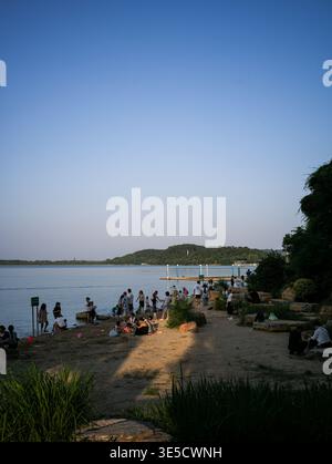 Wuhan, Cina - 16 giugno 2025: Persone che camminano e si rilassano su una spiaggia sabbiosa vicino a un lago calmo circondato da alberi e rocce. L'immagine verticale è lunga Foto Stock