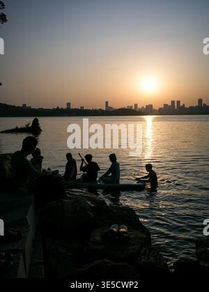 Wuhan, Cina - 16 giugno 2025: Sagome di persone sedute su paddleboard e rocce in un lago con il sole che tramonta su un lontano skyline della città. Il Foto Stock