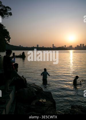 Wuhan, Cina - 16 giugno 2025: Sagome di persone in piedi nell'acqua del lago e sedute sulle rocce sulla riva al tramonto. L'immagine visualizza il bri Foto Stock