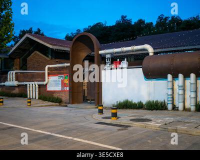 Wuhan, Cina - 18 giugno 2025: Edificio industriale in mattoni rossi ristrutturato con grandi tubi bianchi e un'entrata ad arco al crepuscolo. L'immagine mostra una p Foto Stock