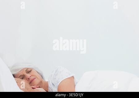 Donna anziana che riposa su un lato della camera da letto con pjs floreali, piumino bianco visibile, spazio fotocopie. Sereno, tranquillo, minimalista, pastello, morbido, calma, Foto Stock