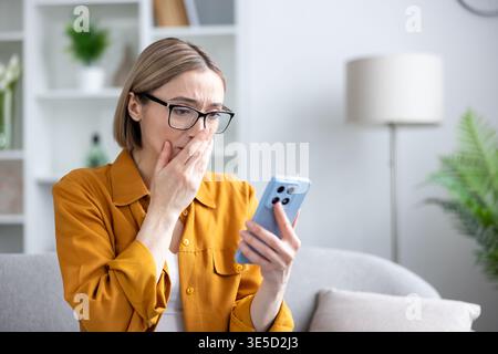 La giovane donna con gli occhiali reagisce con shock e stress mentre legge notizie sconvolgenti sul suo smartphone, coprendo la bocca con incredulità per i contenuti e i messaggi online disturbati Foto Stock
