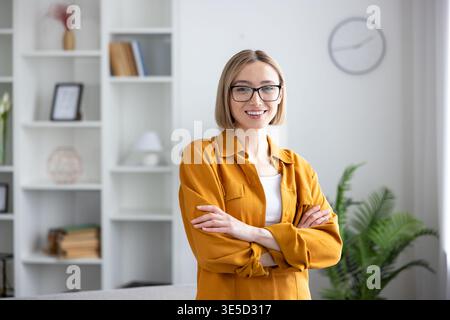 Giovane donna d'affari professionista con occhiali sorridenti alla telecamera, in piedi con le braccia incrociate in un ambiente moderno di casa o ufficio, che rappresenta il successo e la determinazione Foto Stock