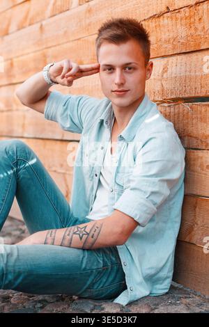 simpatico e affascinante ragazzo alla moda con un'acconciatura in denim con una camicia e un jeans siede a terra vicino a una parete di legno e tiene le dita al suo fianco Foto Stock