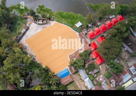 Vista aerea di milioni di grani di riso sono disposti ad asciugare in un mulino mentre i lavoratori li spazzolano con foglie al sole caldo, Dhamrai, Dacca, Bangladesh. Foto Stock