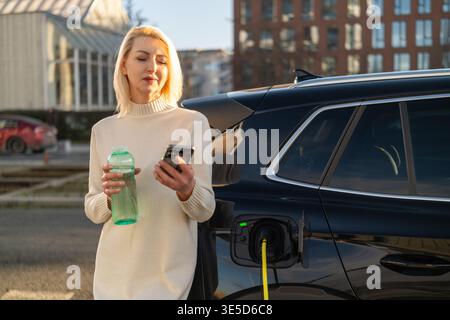 Donna di mezza età che controlla lo smartphone mentre si trova vicino a un veicolo elettrico presso la stazione di ricarica. Luminoso ambiente urbano caratterizzato da un edificio moderno e auto parcheggiate sullo sfondo. Foto Stock
