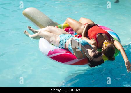 Rilassanti amiche diverse che prendono il sole nella piscina all'aperto, con galleggiante a turbolenza arcobaleno e occhiali da sole Foto Stock