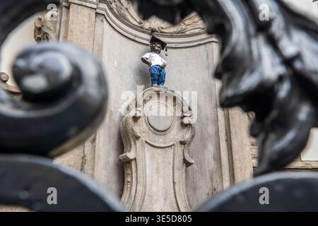 Una vista creativa del Manneken Pis di Bruxelles, incorniciata dai rotoli sfocati di un cancello di ferro. La famosa statua indossa un costume di graduazione con una A. Foto Stock