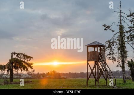 La torre di avvistamento in legno sorge in un prato rurale all'alba, emettendo una calda luce tra i campi e gli alberi lontani. Tranquillo scenario di campagna con un cielo spettacolare, Foto Stock