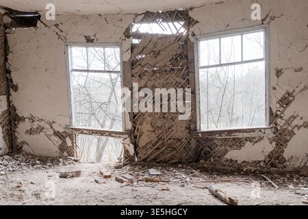 L'interno desolato dell'edificio in rovina presenta finestre frantumate, intonaco sbriciolato, pavimento in detriti e reticolo di legno esposto contro i desolati alberi invernali Foto Stock
