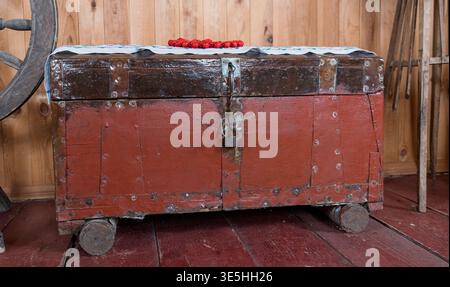 Antica cassa in legno rosso con accessori in metallo e lucchetto appoggiato su ruote in tronchi all'interno di un fienile rustico vicino alla ruota rotante e agli attrezzi agricoli Foto Stock