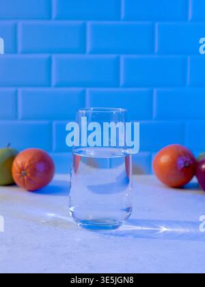 Bicchiere di acqua pulita con luce blu colorata e frutta fresca sul bancone della cucina Foto Stock
