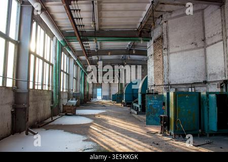 Sala industriale abbandonata, luce del sole che scorre attraverso finestre polverose su un pavimento polveroso, vecchi macchinari abbandonati, rivelando decadimento e negl Foto Stock