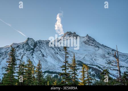 Una montagna con una nuvola di fumo che sale da essa. Il cielo è limpido e blu. La montagna è coperta di neve Foto Stock