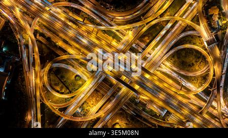 Vista dall'alto verso il basso di un complesso svincolo autostradale di notte. L'esposizione lunga cattura le tracce luminose dei veicoli. Rappresenta la connettività urbana. Foto Stock