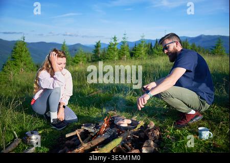 Giovane donna e barbuto accanto al fuoco in un prato di montagna. Una turista femminile sembra rilassata, mentre l'uomo, con gli occhiali da sole, tende a calpestare il fuoco. Vegetazione lussureggiante e montagne lontane sotto il cielo azzurro. Foto Stock