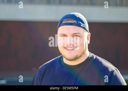 Ritratto di un giovane sorridente che indossa un cappellino da baseball all'indietro e una felpa scura all'aperto, in piena luce naturale. Foto Stock