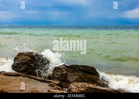 Le potenti onde del lago Issyk-Kul si infrangono contro rocce rocciose lungo una costa rocciosa. I cieli parzialmente nuvolosi migliorano l'atmosfera dinamica e naturale della scena. Foto Stock