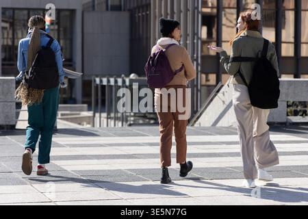 Diverse studentesse che camminano attraverso il campus portando con sé longboard e zaini con foulard Foto Stock