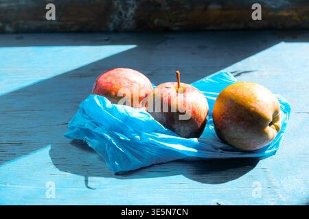 Mele rosse e gialle vivaci poste su un sacchetto di plastica blu su un tavolo in legno dipinto, illuminate da una forte luce naturale della finestra che crea ombre profonde Foto Stock