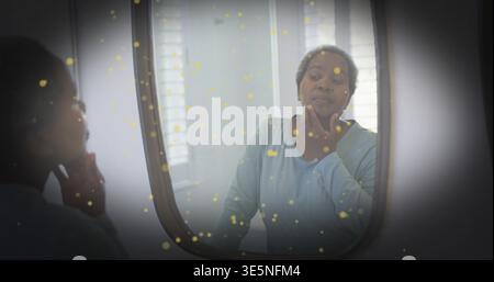 Toccando il mento, donna matura in cima blu che controlla il viso allo specchio del bagno con finestra, macchie gialle Foto Stock