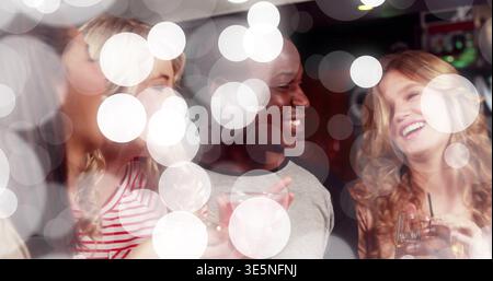 Un gruppo ridendo di quattro amici che chiacchierano e tengono un drink nella sala illuminata, con bokeh circolare Foto Stock