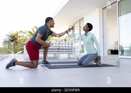 Diverse coppie che toccano gli smartphone mentre si inginocchiano sui tappetini da yoga sulla terrazza di casa, con una bottiglia d'acqua Foto Stock