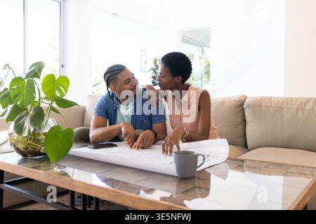 Coppie diverse che rivedono il progetto sul divano in un luminoso soggiorno, con piante in vaso e smartphone Foto Stock