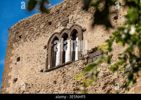 Sessa Aurunca - Campania - Italia - 08-29-2025: Facciata in pietra grezza con triplo arco finestra incorniciata da colonne intagliate, illuminata dal sole sul cielo blu e delicatamente contornata da foglie verdi sul lato. Foto Stock