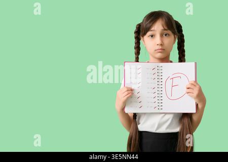 Una studentessa triste che tiene in mano un taccuino con risultati negativi dei test scolastici su sfondo verde Foto Stock