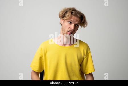 Giovane biondo che fa una faccia sciocca e gli rimbalza la lingua, esprimendo un'emozione umoristica, giocosa e infantile mentre guarda di lato Foto Stock