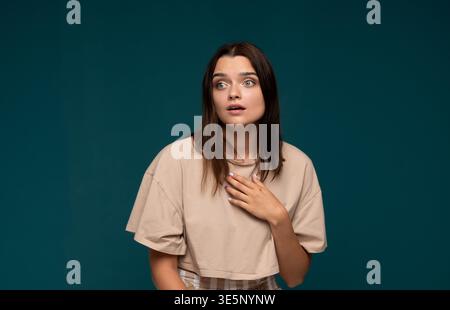 Giovane donna che si sente sorpresa e scioccata, esprimendo incredulità con la mano sul petto sullo sfondo di uno studio Foto Stock