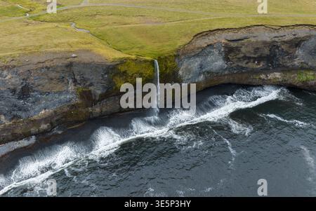 Vista aerea di una cascata che scende da un'aspra scogliera nel mare turbolento, le onde spumeggianti che si infrangono contro le rocce scure sottostanti, Húnabyggð, IC Foto Stock