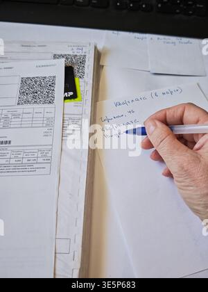 Kirov, Russia - 18 marzo 2026: Una persona scrive appunti mentre è circondata da bollette e documenti finanziari su una scrivania a casa, concentrandosi sulla casa Foto Stock