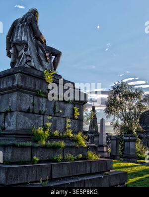 Una suggestiva necropoli al crepuscolo, caratterizzata da una statua intemprata in cima a un monumento in pietra, erba e felci sovrastate, tombe storiche e un umore Foto Stock