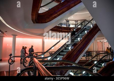 Le persone viaggiano e camminano accanto alle eleganti scale mobili in legno all'interno di Princes Square, un moderno centro commerciale con illuminazione calda, architetto Foto Stock