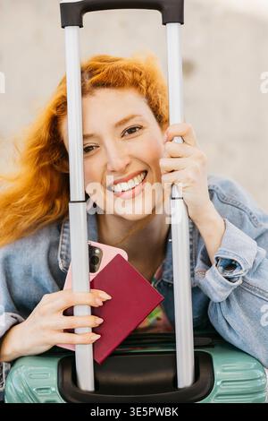 Giovane donna sorridente, con passaporto e telefono in mano, seduta su valigia rotolante Foto Stock