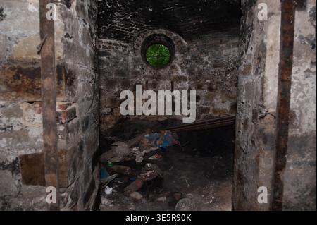 Dungeon in mattoni abbandonati con finestra circolare di colore verde e pavimento impilato con detriti in texture rustica scura che mostrano decadimento e negligenza nel tempo Foto Stock