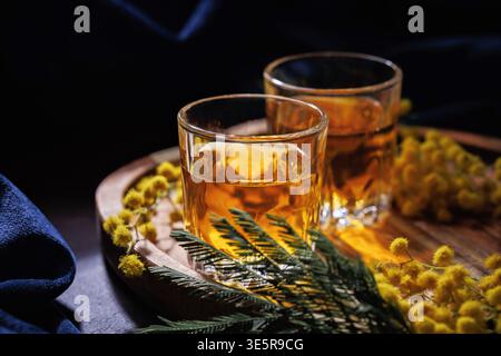 Bicchieri da whisky con fiori di Mimosa su vassoio di legno bevanda calda primaverile natura morta Foto Stock