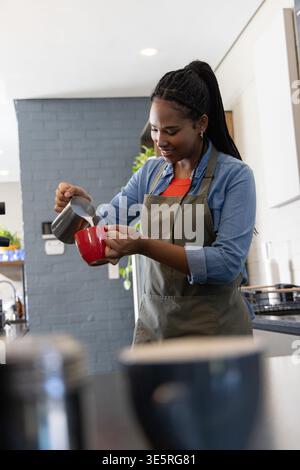 Donna afroamericana che versa latte dalla caraffa di metallo nella tazza rossa dietro il bancone con grembiule Foto Stock