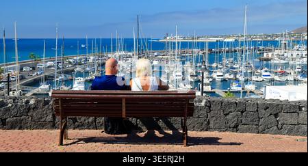 Coppia matura seduta su una panchina che si affaccia su Marina Rubicon, godendo della vista sul mare, Playa Blanca, Lanzarote, Isole Canarie, Spagna Foto Stock