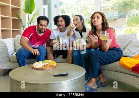 Quattro amici seduti su un divano sezionale nel soggiorno, guardano lo schermo e condividono spuntini e birra Foto Stock
