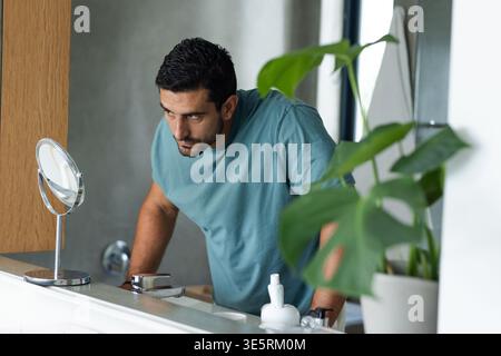 Uomo con il barbuto che indossa una T-shirt color tè appoggiata sul lavandino del bagno, guardando lo specchio mentre si preme il bancone Foto Stock