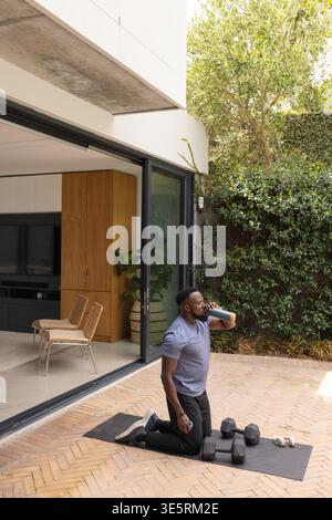 Uomo afro-americano inginocchiato sul tappetino del patio con abbigliamento sportivo, acqua potabile e auricolari con pesi nelle vicinanze Foto Stock