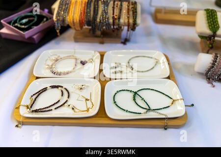 Vari bracciali e collane con perline esposti su piatti in ceramica bianca e banchi in legno. Foto Stock