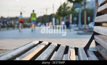 Concentratevi su una panchina di legno, gente offuscata che corre e si rilassa in un parco cittadino all'ora d'oro Foto Stock