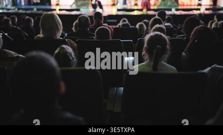 Persone sedute in fila, che si godono uno spettacolo. Il pubblico guarda il palco in un auditorium poco illuminato Foto Stock