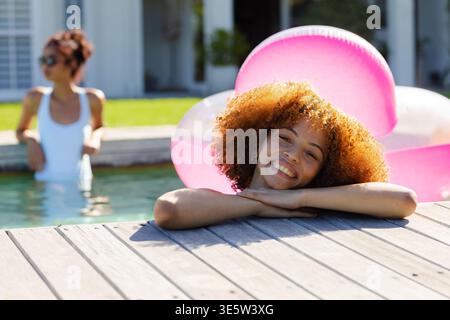 Amiche afroamericane che ridono e si appoggiano sul ponte della piscina in legno con galleggiante rosa e sfumature Foto Stock